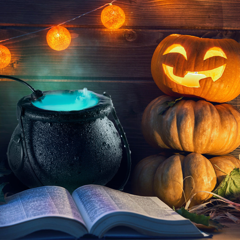 Black Halloween cauldron emitting blue-lit mist beside pumpkins and an open book